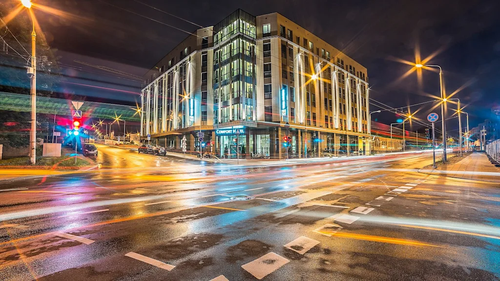 En moderne hotelbygning med oplyste vinduer står på et gadehjørne om natten, omgivet af lysstriber fra passerende køretøjer. Tekst: "Comfort Hotel."