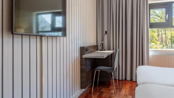 A modern workspace with a wall-mounted television above, situated beside a large window with curtains, in a brightly lit room.