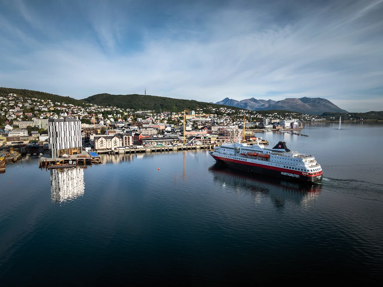 Ett stort passagerarfartyg seglar genom en hamn med en stad inbäddad mot kullar och berg under en blå himmel.