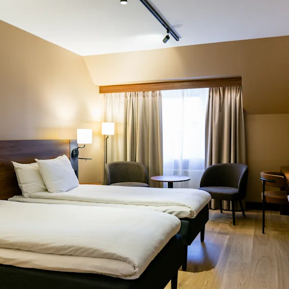 Mysigt hotellrum med två enkelsängar, fåtöljer vid fönstret och varm belysning.