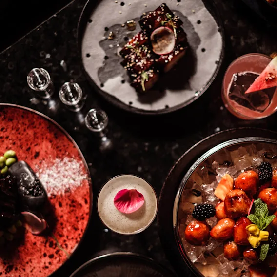 Assorterte gourmetretter på mørkt bord; en cocktail med en blomst, frukt på is og diverse anrettede måltider. Dempet belysning fremhever den elegante presentasjonen.