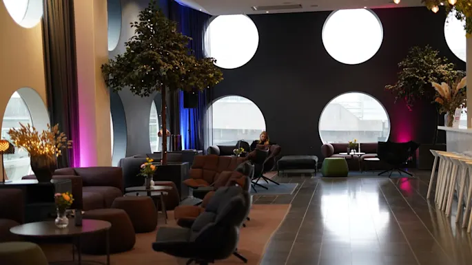 Comfortable seating in a lounge area; a person reads on a sofa. Large circular windows and indoor trees add ambiance, while tables hold vases of flowers. 