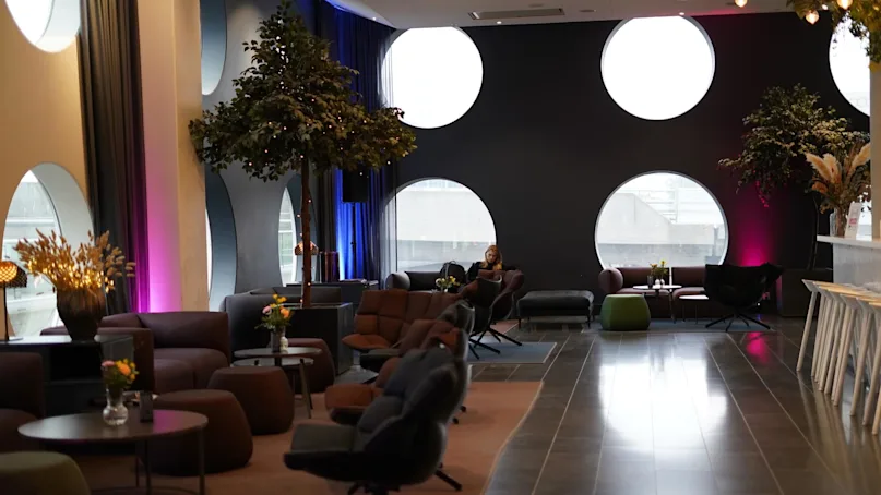 Comfortable seating in a lounge area; a person reads on a sofa. Large circular windows and indoor trees add ambiance, while tables hold vases of flowers. 