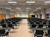Rækker af sorte stole og skriveborde er arrangeret mod en stor hvid skærm i et rummeligt konferencelokale med gitterloft og brunt tæppe.