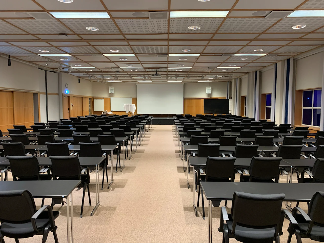Rows of black chairs and desks are arranged facing a large white screen in a spacious conference room with a grid ceiling and brown carpet.