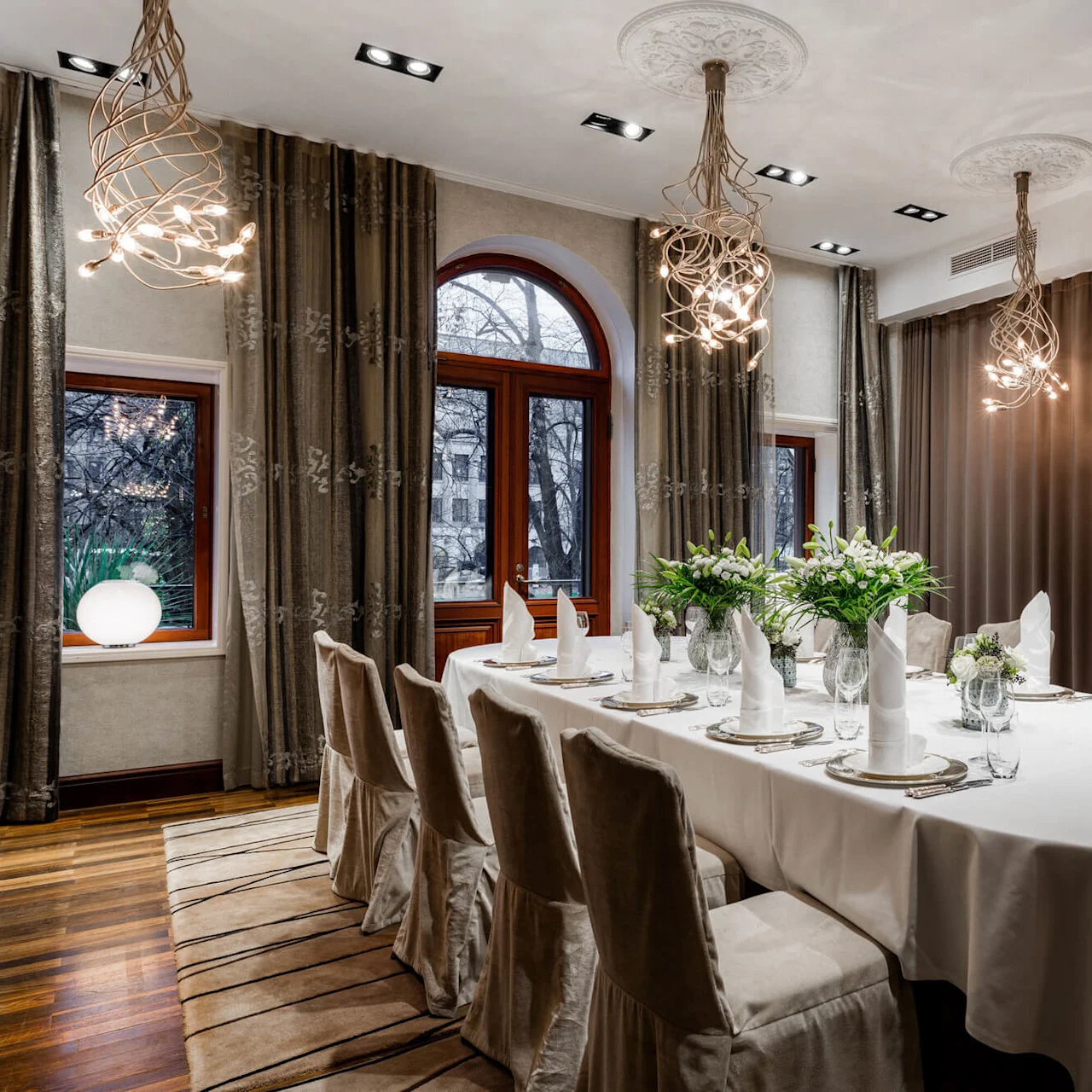 An elegant dining table set with white napkins and flowers, surrounded by beige chairs, under ornate chandeliers, in a room with draped windows.