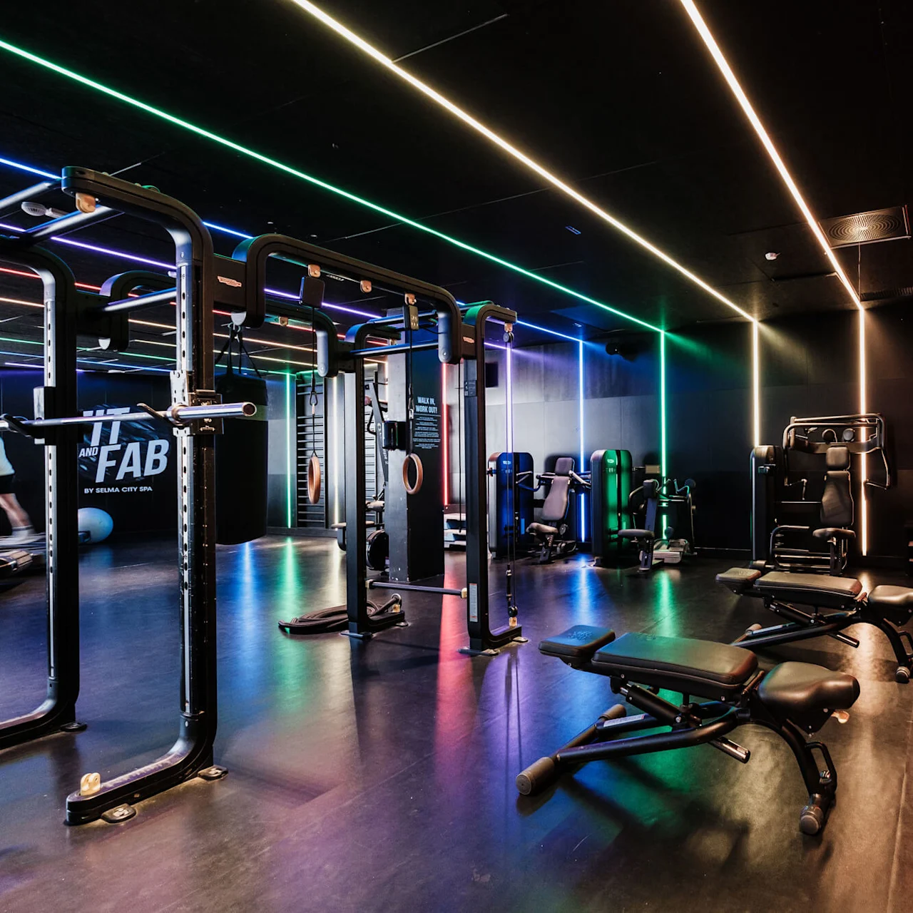 Gymutrustning är arrangerad i ett svagt upplyst rum med färgglada LED-lampor i taket. Bänkar, vikträcken och maskiner fyller utrymmet. Synlig text: "FIT AND FAB BY SELMA CITY SPA."