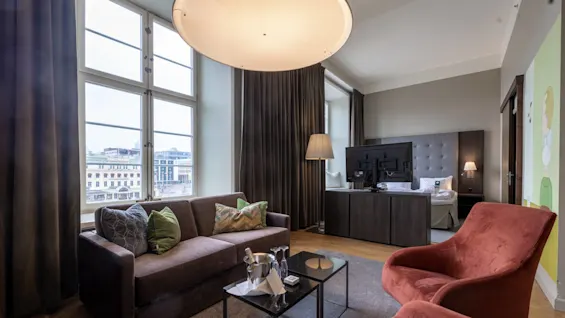 A luxurious hotel room features a sofa and two chairs around a coffee table, with champagne. Large windows overlook a cityscape, and a bed is partially visible behind a stylish divider.
