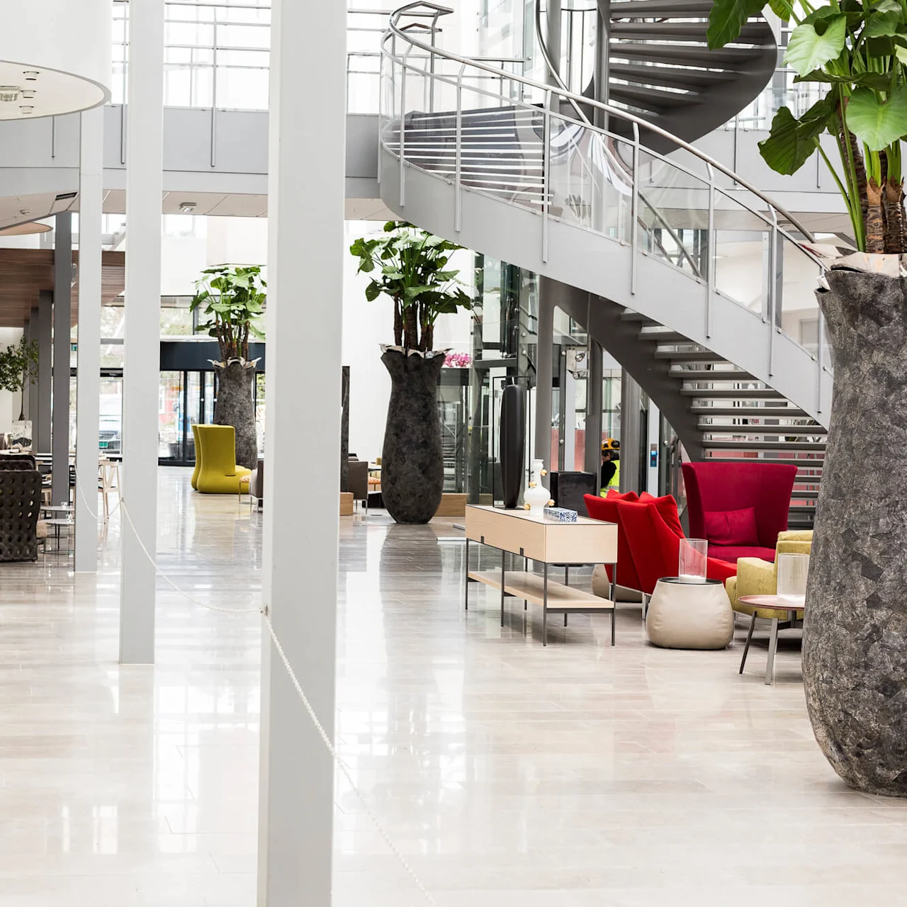 Store vaser med planter dekorerer et moderne atrium med en elegant spiraltrapp og fargerike sittearrangementer. Lys naturlig belysning filtreres gjennom store vinduer, og skaper en romslig, moderne stemning.