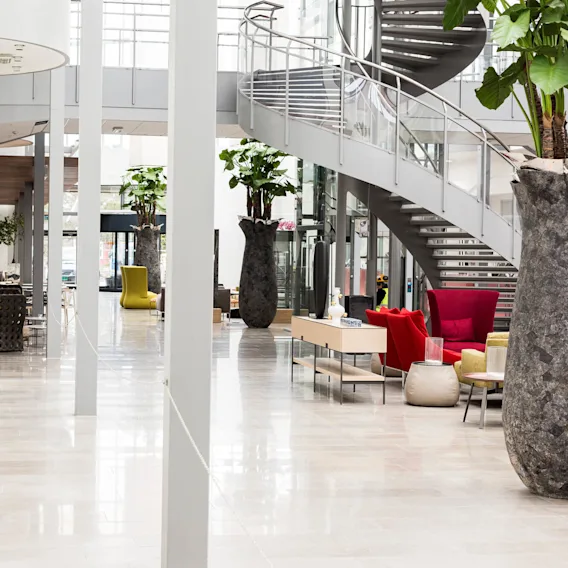 Store vaser med planter dekorerer et moderne atrium med en elegant spiraltrapp og fargerike sittearrangementer. Lys naturlig belysning filtreres gjennom store vinduer, og skaper en romslig, moderne stemning.
