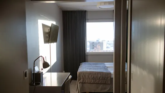 A small, tidy bedroom features a neatly made bed, a wall-mounted TV, and a desk with a chair. Sunlight streams through a window with dark curtains, revealing a distant city view.