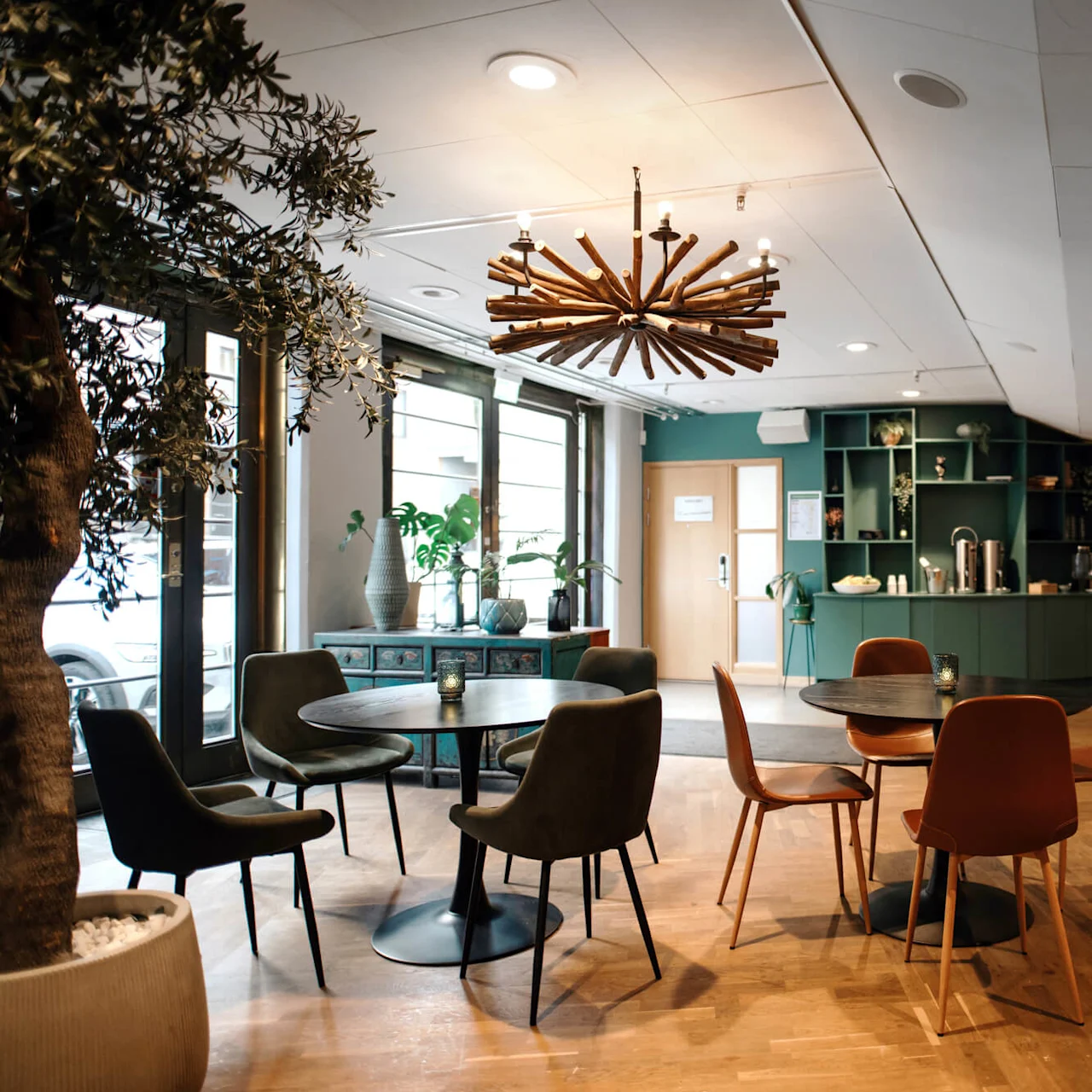 A modern office lounge with tables, chairs, and decorative lighting, woods floors and green shelving units.