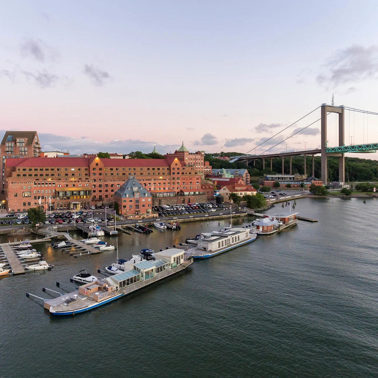 Boats docked on a marina beside a red-roofed building, surrounded by parked cars. A large suspension bridge spans over water in the background, with greenery visible onshore.
