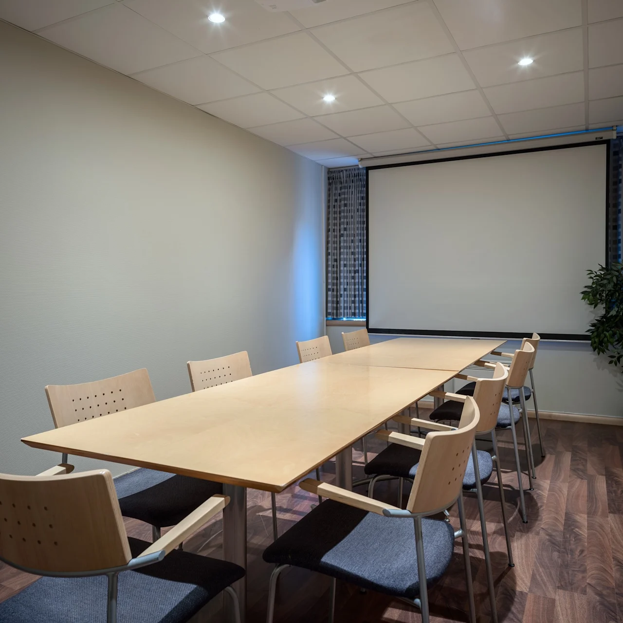 Ett långt träbord med åtta stolar omger det, beläget i ett konferensrum med en stor tom skärm, mönstrade gardiner och en krukväxt mot en beige vägg.
