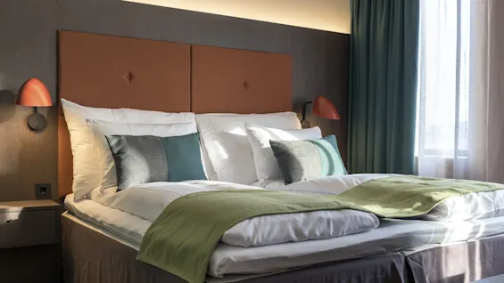 A neatly made bed with white sheets and green accents stands in a modern, well-lit bedroom featuring red lamps and a window with curtains.
