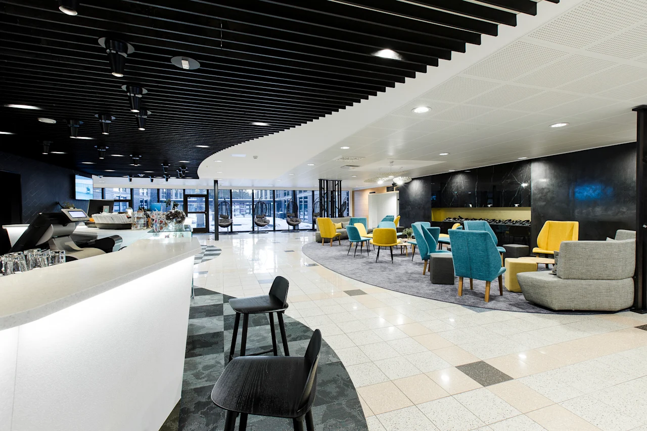 Valoisa, moderni lounge, jossa värikkäät keltaiset ja siniset tuolit matalien pöytien ympärillä matolla, vierekkäin tyylikkään valkoisen baaritiskin kanssa, tumman katon alla.