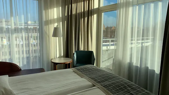 Valoisa hotellihuone, jossa on mukava sänky, pieni oleskelutila ja suuret ikkunat kaupunkinäköalalla.