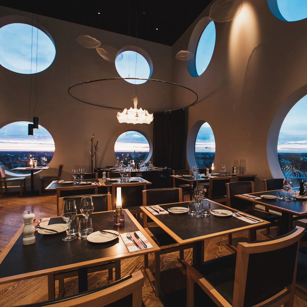 Borde med dækket spisning har udsigt over byens skyline gennem store cirkulære vinduer i en stilfuld restaurant med trægulv og elegant belysning.