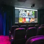 En lille biograf med røde stole vender mod en stor skærm med filmvalg. Dæmpet lilla belysning og mørke gardiner skaber en intim atmosfære.