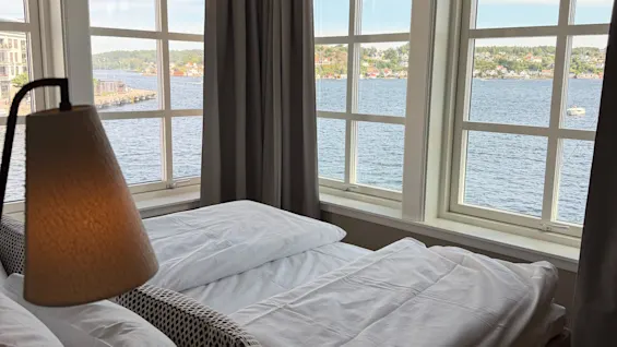A cozy bedroom with a bed and a lamp, offering a beautiful view of a calm lake and distant land through large windows.