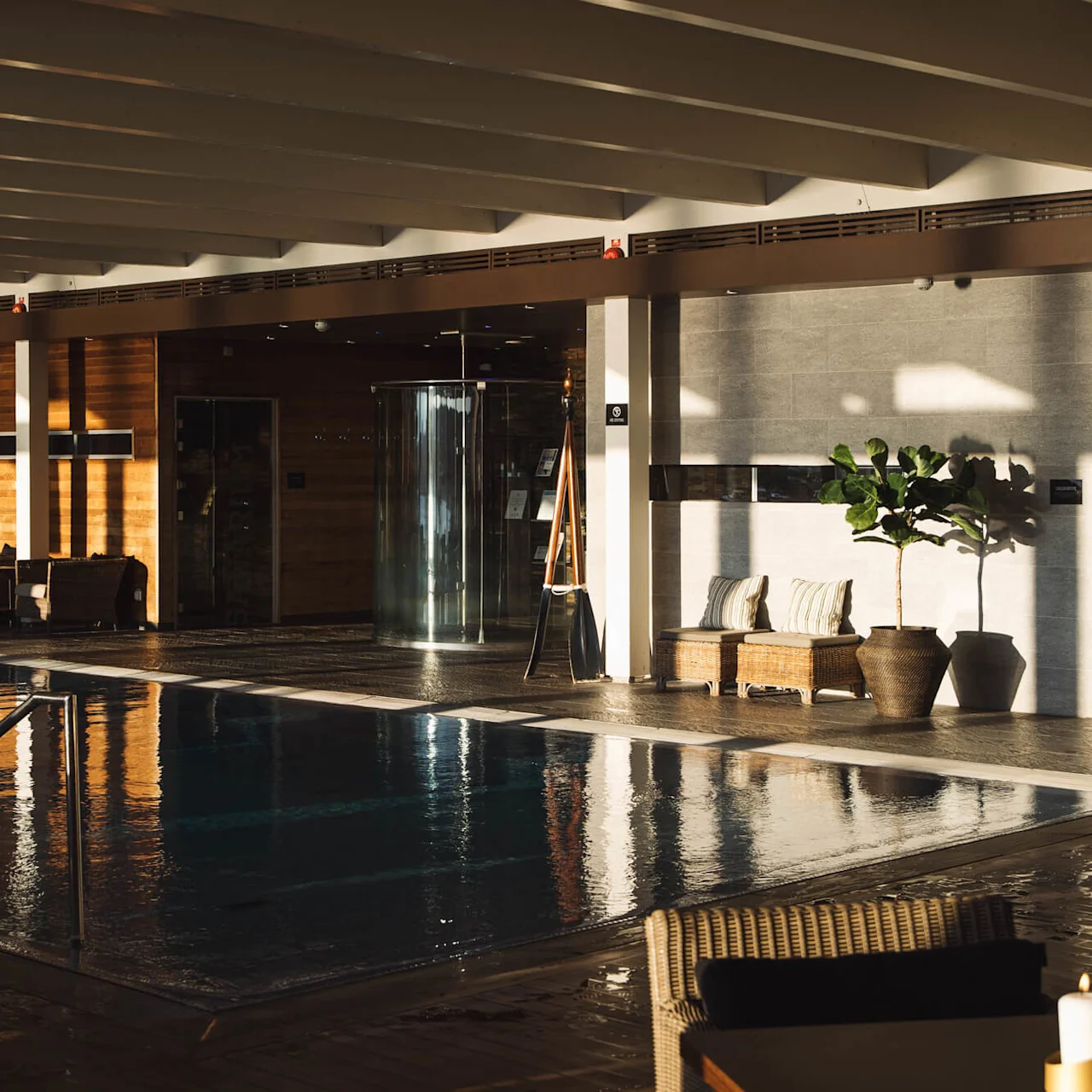 Swimming pool reflects sunlight, surrounded by lounge chairs and potted plants. Warm wooden panels and cushioned seating create a serene, indoor relaxation space.