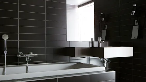 Et moderne baderom har en rektangulær vask og badekar, med elegante, mørke flislagte vegger og kromfester, ved siden av et stort vindu som skaper en minimalistisk og moderne atmosfære.
