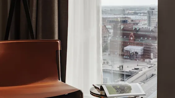 Et koselig hotellrom med en brun skinnstol, en gulvlampe og et magasin på et sidebord, med utsikt over byen.