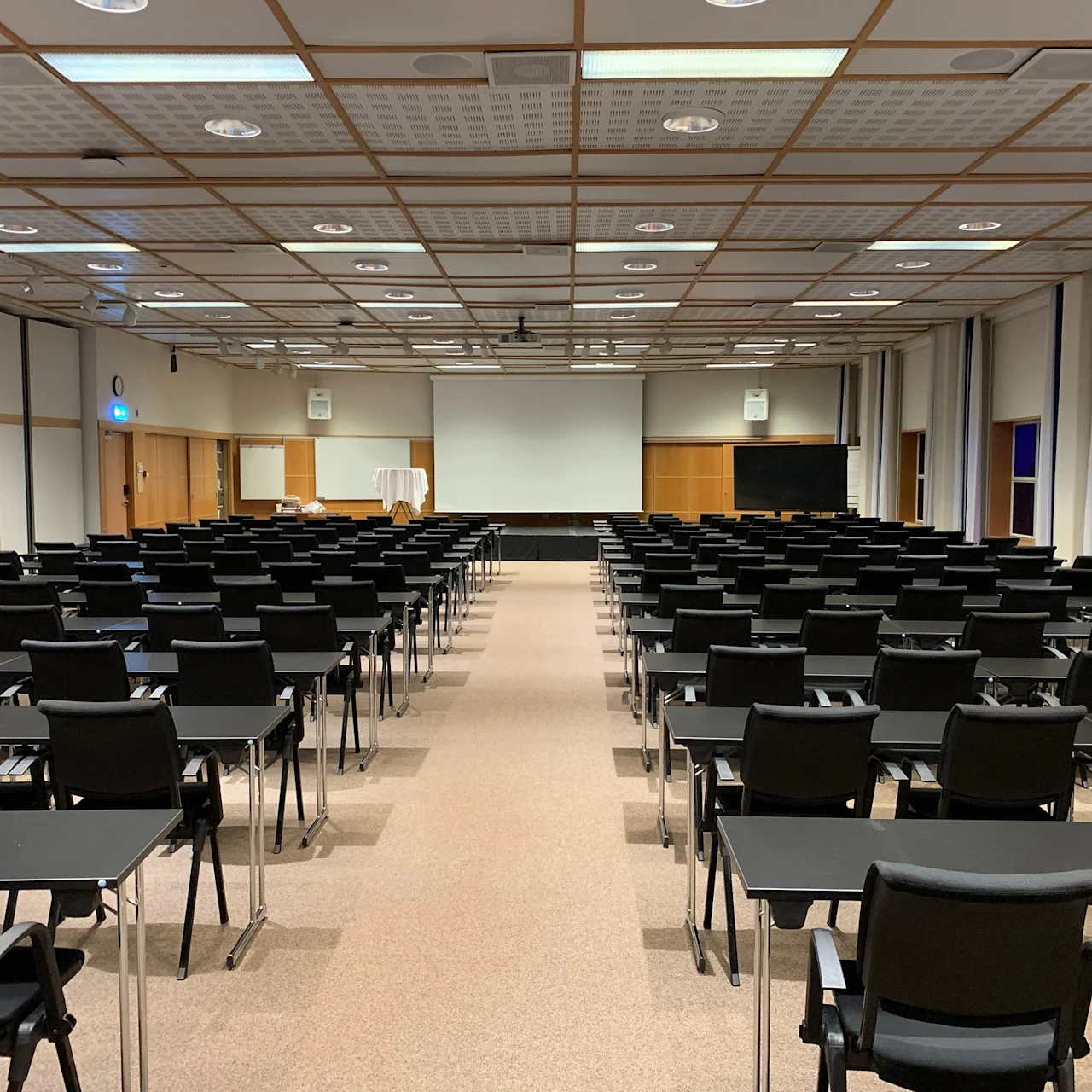 Rows of black chairs and desks are arranged facing a large white screen in a spacious conference room with a grid ceiling and brown carpet.