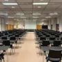 Rows of black chairs and desks are arranged facing a large white screen in a spacious conference room with a grid ceiling and brown carpet.