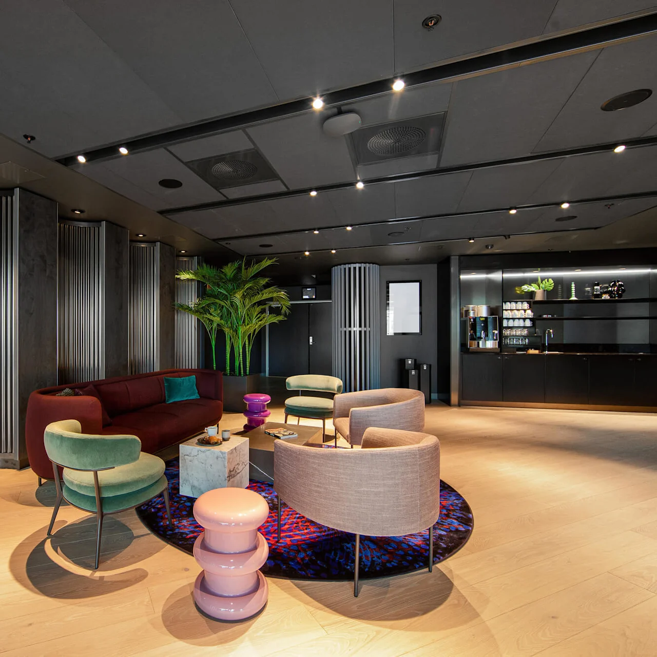 A modern lounge features stylish chairs, a burgundy sofa, and a circular rug. A potted plant decorates the space, and a beverage station is visible in the background.