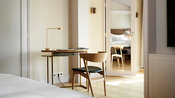 Et skrivebord med lampe og stol står i et minimalistisk, godt opplyst soverom. Et speil på døren reflekterer en seng omgitt av hvite vegger og lys tregulv.
