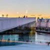 A modern building with a sloped marble roof features people walking against a sunset sky. Reflections appear in large windows, with a waterfront and city buildings in the background.
