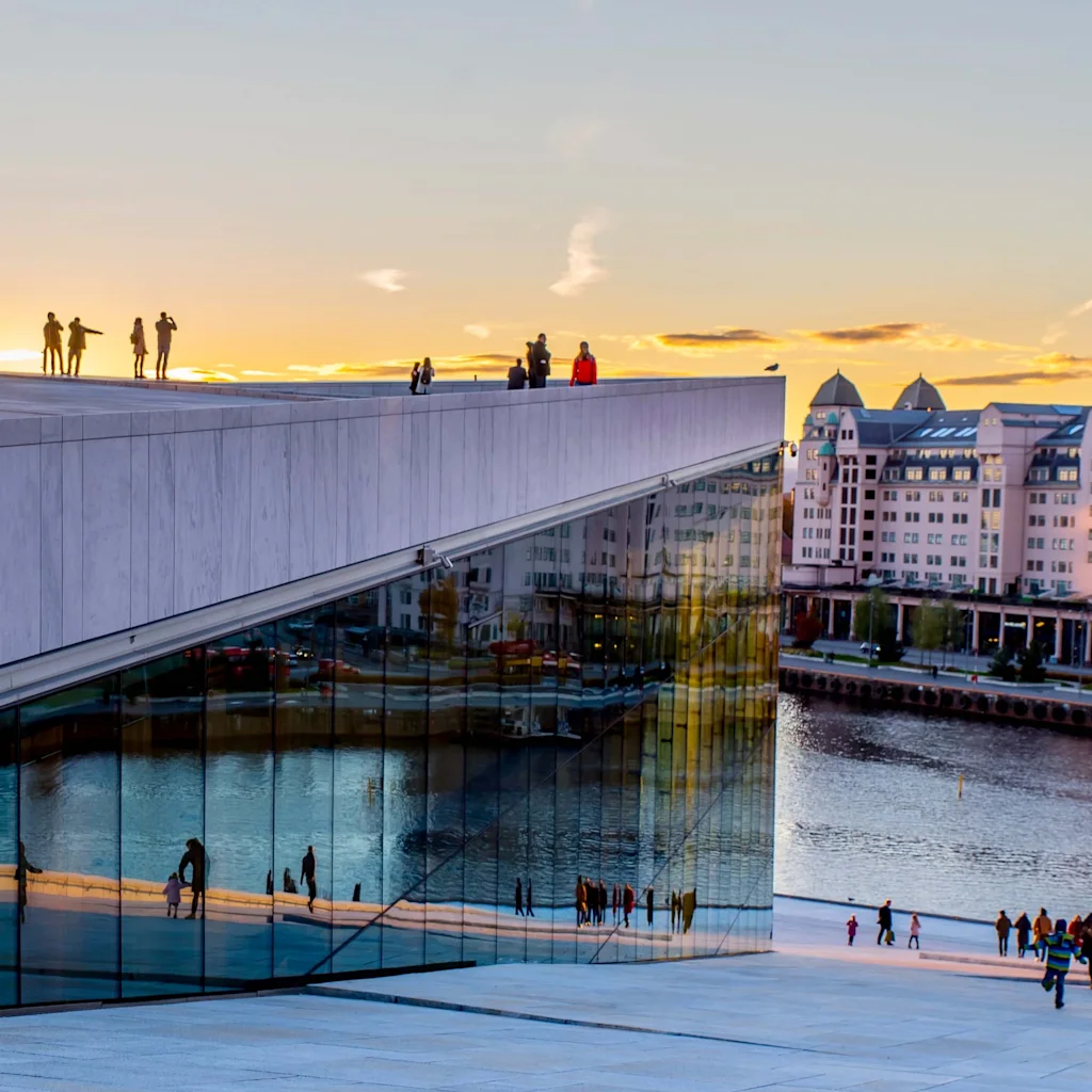 A modern building with a sloped marble roof features people walking against a sunset sky. Reflections appear in large windows, with a waterfront and city buildings in the background.