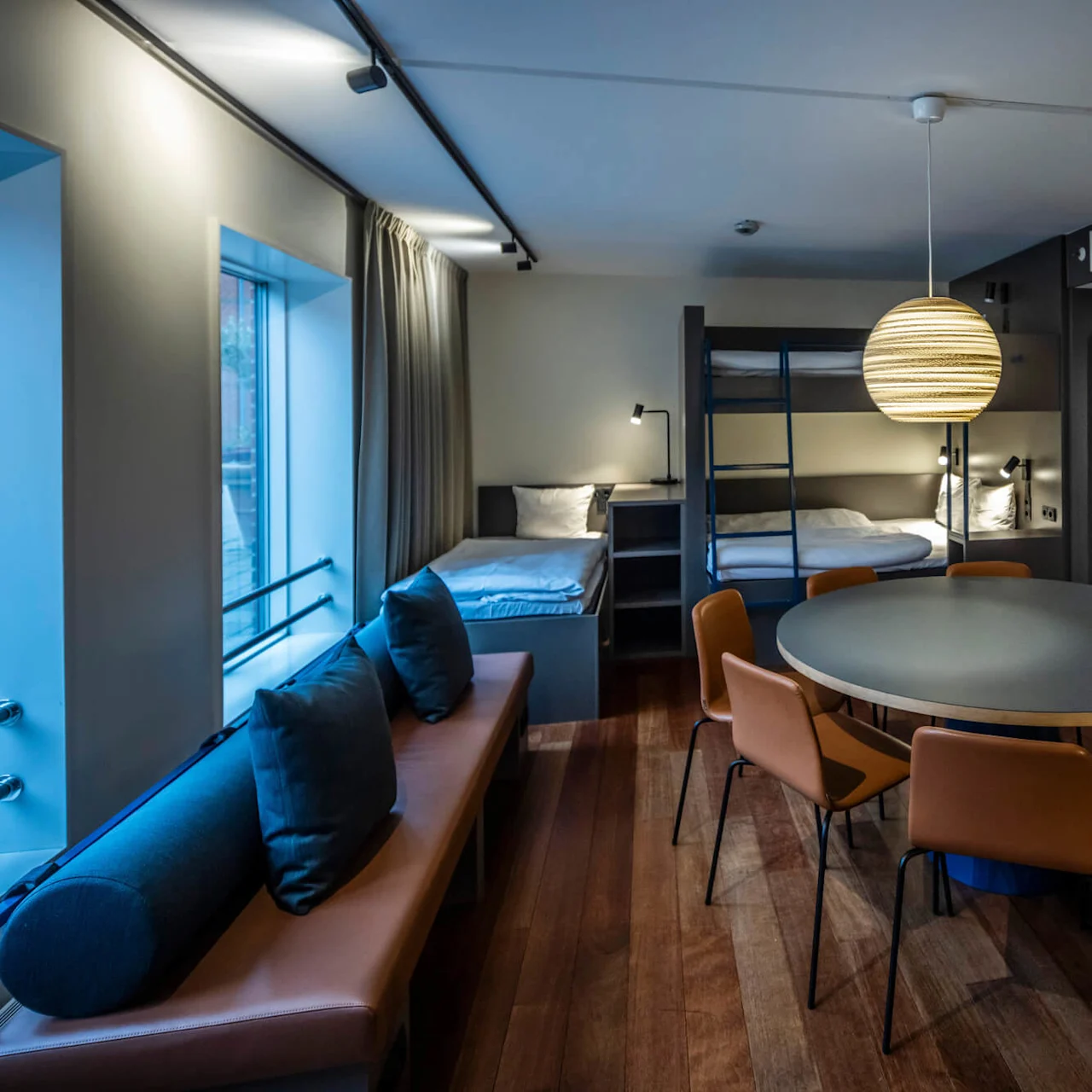 A large hotel room with bunk beds, a single bed, a round dining table with orange chairs, and a seating bench with blue cushions. Large windows provide natural light.