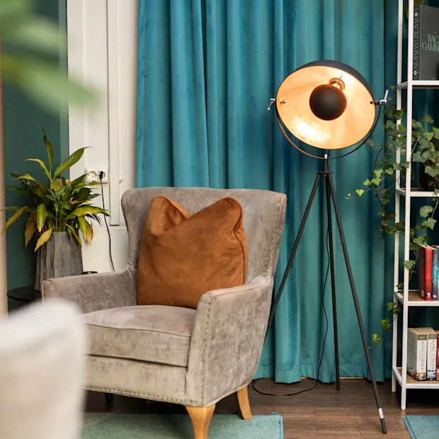 Lænestol med brun pude står i rum med blå gardiner. Lampe på stativ står ved siden af. Reol med bøger og dekorative genstande står i hjørnet. Frodig plante komplementerer det hyggelige opstilling.