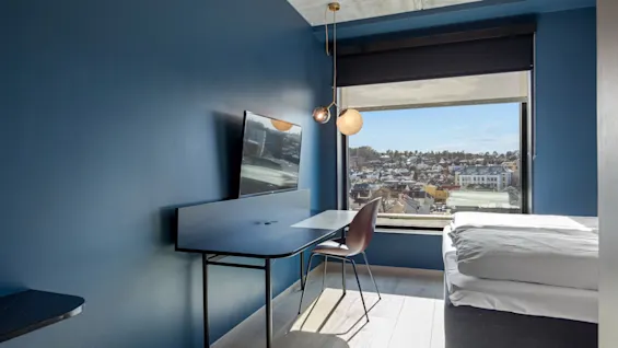 Et moderne hotellrom med blå vegger, et skrivebord, stol og TV, ved siden av en seng og et stort vindu med utsikt over en by.