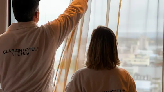 Två personer i badrockar som tittar ut genom ett hotellfönster på utsikten över staden.