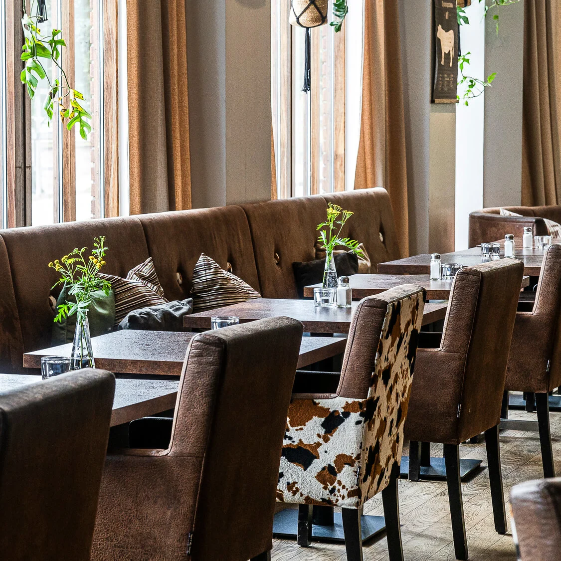 Elegant inredd restaurang med bruna stolar, bord och dekorativa kuddar, nära höga fönster med tunga gardiner.