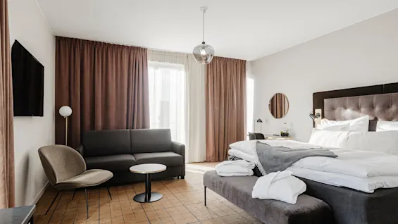 Et moderne hotelværelse har en seng med hvidt sengetøj, en grå sofa, et rundt bord, en stol og beige gardiner fra gulv til loft. Et TV er monteret på væggen.