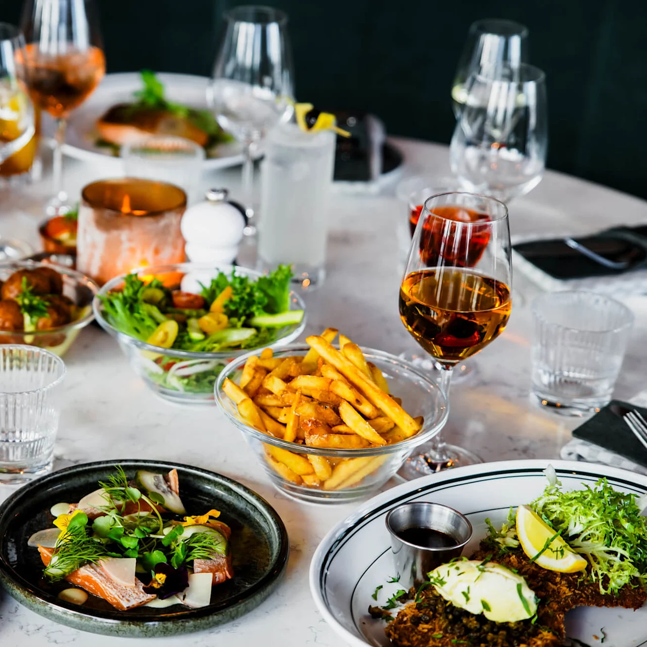 Forskellige retter og drikkevarer arrangeret på et hvidt marmorbord, med pommes frites, salater, skaldyr og vinglas. Omgivelserne antyder en finere middagoplevelse med elegant borddækning.