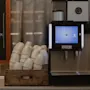 En kaffemaskin visar en meny bredvid en låda med staplade koppar på en bänk. I närheten står en griffeltavla med texten: "Har du mer mat i magen? Det kan du med sig fråga receperionen."