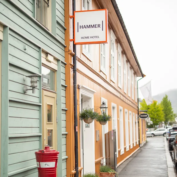 Bygning skilt med teksten "HAMMER HOME HOTEL" på en oransje bygning. Toetasjes bygninger langs en våt gate med parkerte biler. Trær synlige i bakgrunnen.