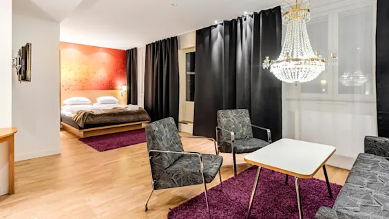 Et romslig hotellrom har en seng med et brunt teppe, en krystallkrone og et sitteområde med to stoler og et bord over et lilla teppe.