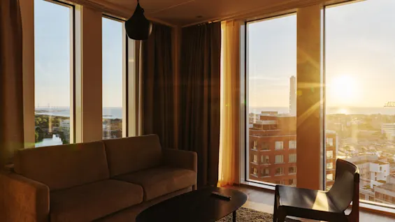 En koselig stue med sofa og stol, badet i den varme gløden fra en solnedgang over en bysilhuett.