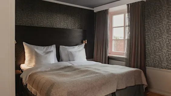Ett välkomnande hotellrum på Home Hotel Grand, Helsingborg, med en prydligt bäddad säng, mörka mönstrade väggar och ett fönster.