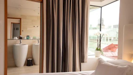 Ett hotellrum med en säng, ett modernt badrum med två handfat och ett fönster med utsikt över staden med en orkidé.