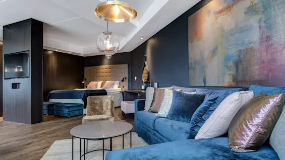 Moderni ja tyylikäs hotellihuone, jossa on sininen sohvaryhmä ja taustalla näkyvä parivuode.