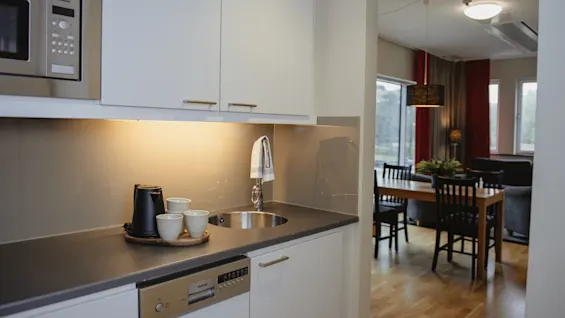 Kitchen counter with a sink holds a kettle and cups. Bright, modern dining area with wooden table and black chairs is visible in the background, near large windows.