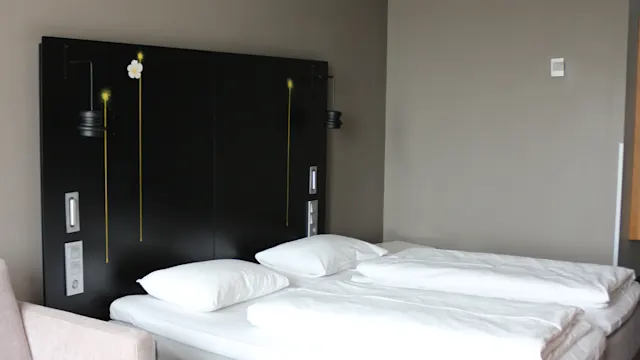 En dubbelsäng med vita sängkläder står mot en mörk sänggavel med enkla blommönster. En mönstrad blå matta och beige soffa ramar in det moderna, minimalistiska rummet.