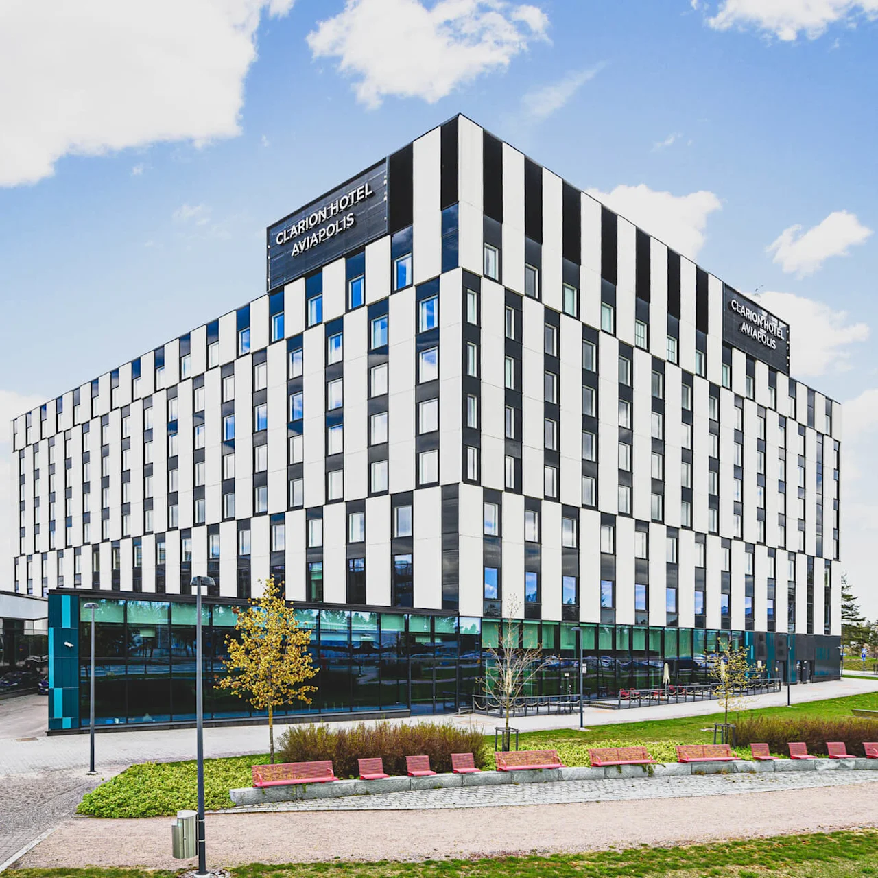 A modern, multi-story building with a black and white facade labeled "CLARION HOTEL AVIAPOLIS" stands amidst a landscaped area with benches on a clear day.